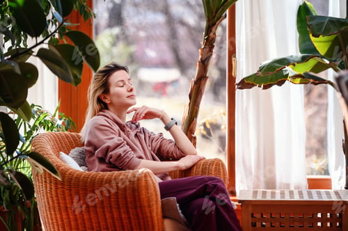 Preview: Young blonde woman sits in rattan armchair in pink-violet clothes in front of window