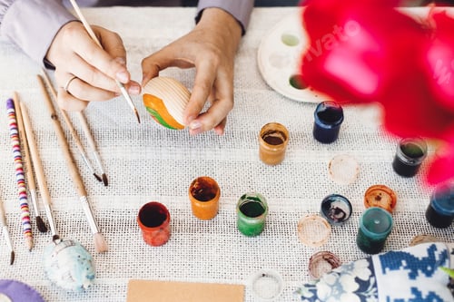 Preview: Hands holding and painting easter egg on rustic table with paint, brushes, tulips bouquet