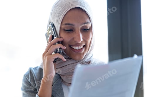 Preview: Muslim business woman wearing hijab talking with her smart phone while holding papers in the office.