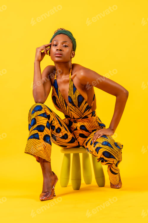 Preview: Young african woman with traditional costume on yellow background, sitting looking at camera