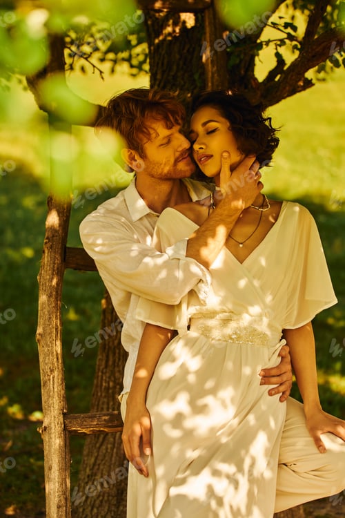 Preview: happy redhead groom embracing tender asian woman in wedding dress under tree outdoors, rural setting