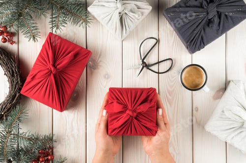 Preview: Female hands hold christmas present in furoshiki package, top view. Spruce branches, scissors, cup