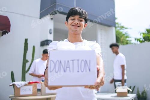 Preview: Asian Young Man Holding a Donation Box Full of Food Supplies at Charity Center