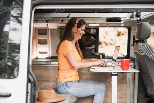 Preview: Mature woman working inside mini van camper with computer laptop wearing wireless headphone