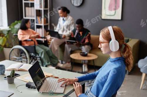 Preview: Red haired young woman wearing headphones in office working with laptop