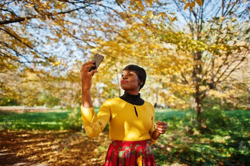 Preview: African american girl at yellow and red dress at golden autumn fall park making selfie on phone.