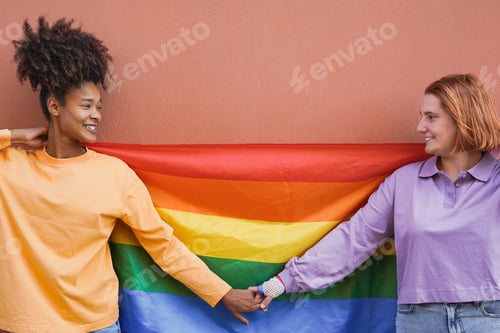 Preview: Young lesbian couple with rainbow flag holding hands outdoor at pride parade