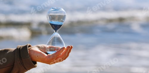 Preview: Time is running fast. Hourglass with blue sand inside in mature woman's hand symbolize the brevity