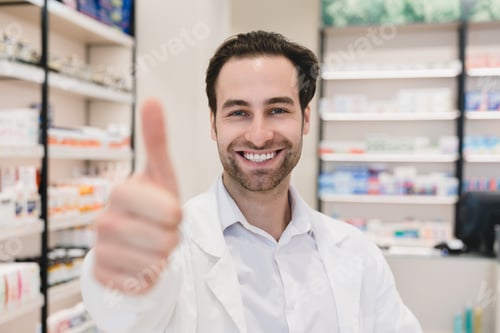 Preview: Pharmacist showing thumb up in camera in white medical coat in pharmacy drugstore