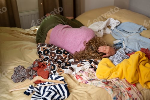 Preview: Woman buried face down in an assortment of colorful clothing on a bed.