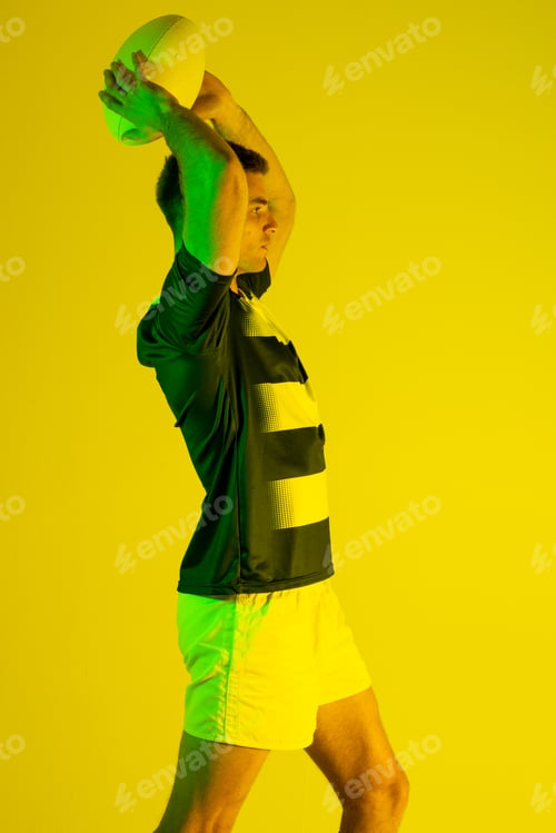Preview: Caucasian male rugby player with rugby ball over yellow lighting