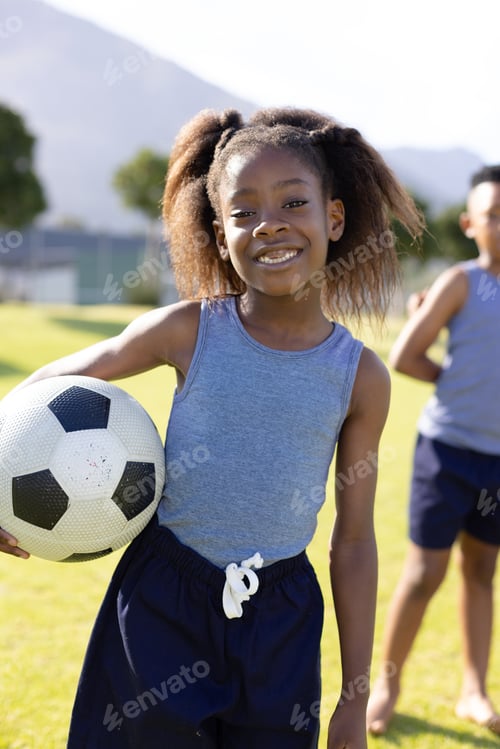 Preview: Portrait of happy african american schoolgirl holding football on field at school