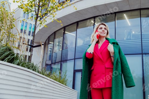 Preview: mature blonde curly woman in suit walks in business district and uses her phone.