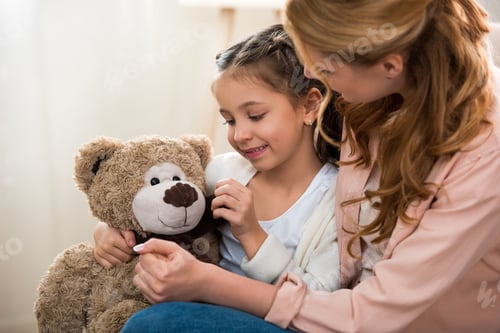Preview: beautiful happy mother and daughter playing with teddy bear at home