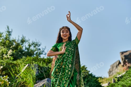 Preview: excited young indian woman in stylish green sari posing in summer park on background