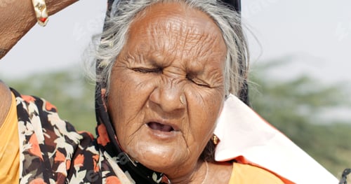 Preview: Closeup of an old South Asian woman with her eyes closed and a Covid mask dangling from one ear