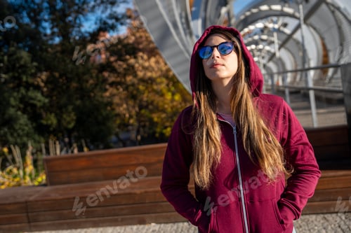 Preview: Young woman wearing sunglasses and hoodie standing on modern bridge