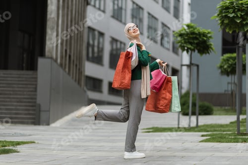 Preview: Muslim woman, wearing grey pants and green jacket, walking at the street with colorful shopping bags