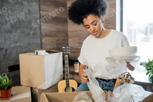 Preview: african american woman unpacking tableware near cardboard box in new house