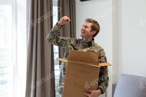 Preview: Smiling man in camouflage holding cardboard box in new apartments