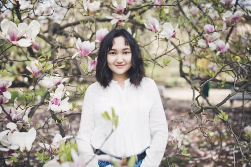 Preview: Cheerful asian young woman in a white blouse under the magnolia blooming tree.