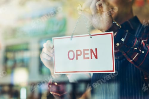 Preview: Opening day has arrived. Closeup shot of a young man hanging up an open sign in a shop window.