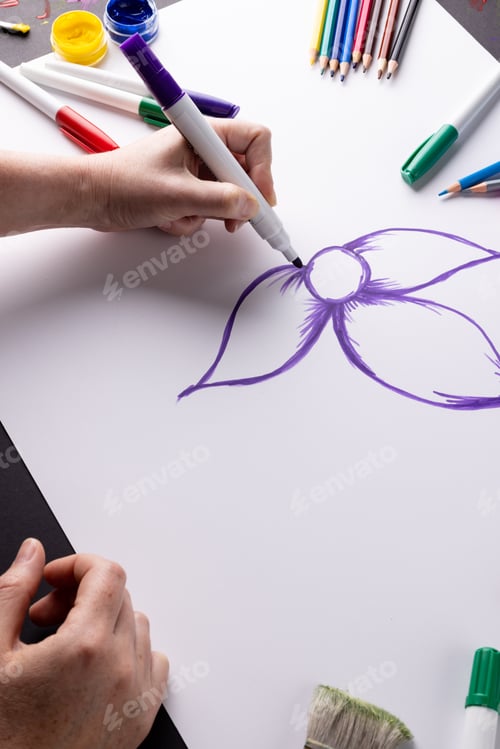 Preview: Vertical image of hands drawing purple flower on paper and art materials on table top, copy space