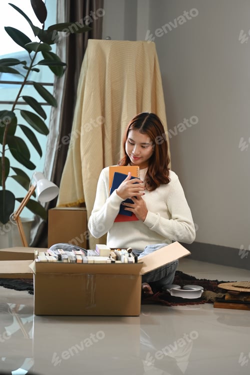 Preview: Happy young woman sitting among various carton boxes with stuff, enjoying moving in new house