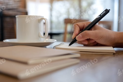 Preview: Closeup image of a hand writing on blank notebook with coffee cup on table in cafe