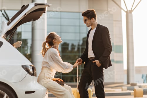 Preview: Handsome man in black suit touches girlfriend hand and smiles. Blonde woman in white blouse, beige