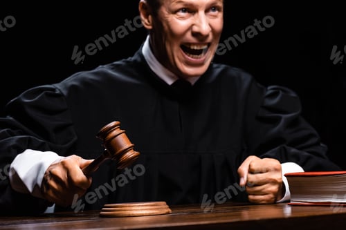 Preview: angry judge in judicial robe sitting at table and hitting with gavel isolated on black