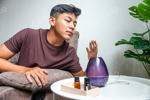 Preview: Young Man Sits on Sofa with Eyes Closed and Enjoys Clean Air Spreading with Modern Humidifier