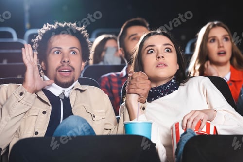 Preview: selective focus of multiethnic and shocked friends holding hands and watching movie in cinema