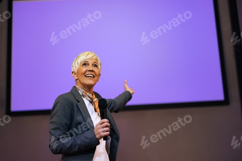 Preview: Senior female holding a microphone, giving a presentation to the public.