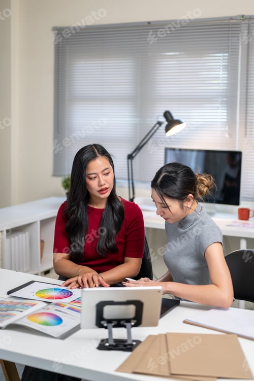 Preview: Asian woman talking to friend coworker over color chart aside tablet at table in home office studio.