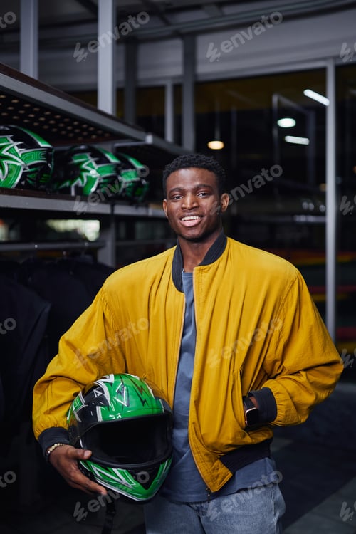 Preview: cheerful african american kart racer in yellow bomber jacket holding helmet, male driver, go-cart