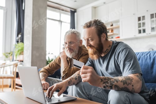 Preview: Smiling gay man holding credit card near mature partner at home