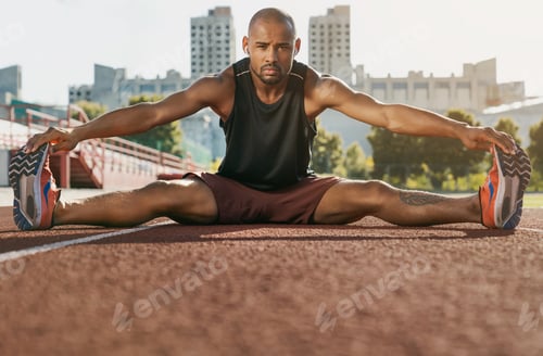 Preview: Sporty muscular african male athlete in earphones stretching legs while sitting at the stadium race