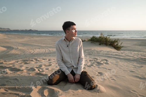 Preview: a young guy sits on the sand in a beige shirt and looks towards the sunset, white patches on his fac