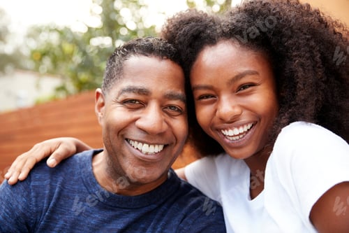 Preview: Middle aged black dad and teenage daughter embracing and smiling to camera, close up