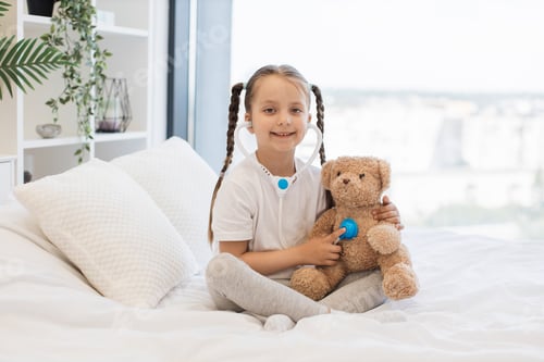 Preview: Pretty kid with toy stethoscope examining teddy bear in bed