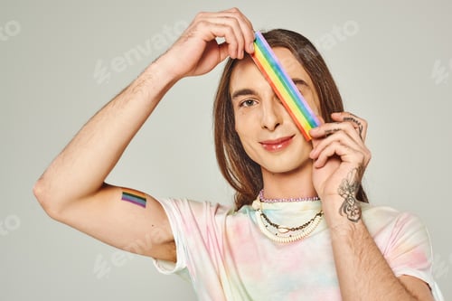 Preview: happy and tattooed gay man with long hair and tie dye t-shirt holding rainbow lgbt flag near face