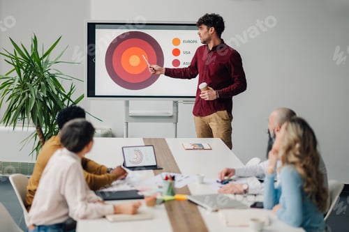 Preview: Young businessman holding takeaway coffee and giving presentation to colleagues in meeting room