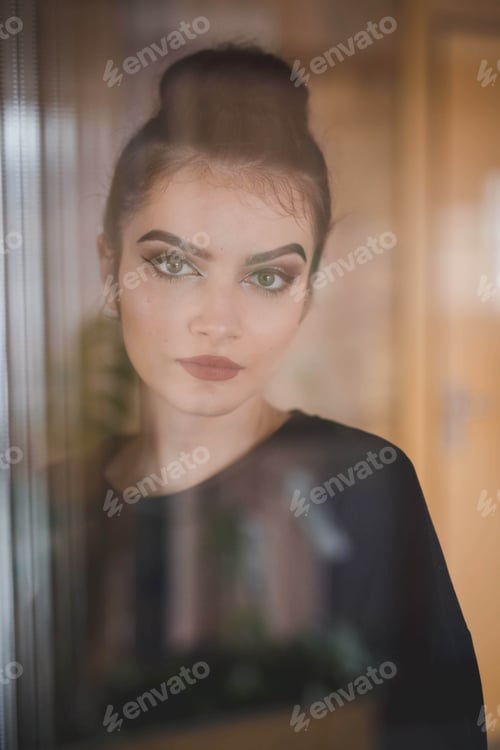 Preview: Young Bosnian woman with a bun hairstyle and makeup looking at the camera through the window