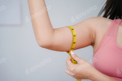 Preview: Woman wearing sportswear is measuring her biceps with a yellow measuring tape