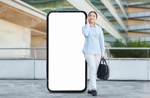 Preview: Woman standing by a large blank phone screen while talking on the phone outdoors