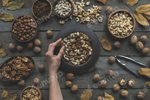 Preview: top view of human hand and various nuts in bowls with dried autumn leaves on table