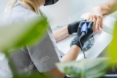 Preview: Female master using electric nail file while doing manicure for client in beauty salon