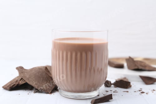 Preview: Tasty chocolate milk in glass and pieces of chocolate on white tiled table, closeup