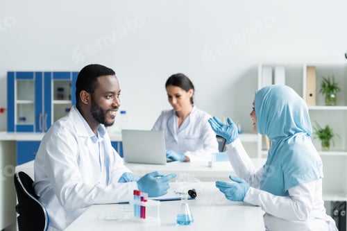 Preview: Smiling multiethnic scientists talking near medical equipment in laboratory